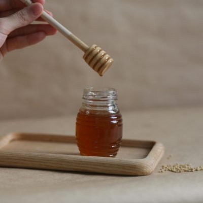 A hand holding a honey dipper over a jar on a wooden tray, showcasing the golden liquid.